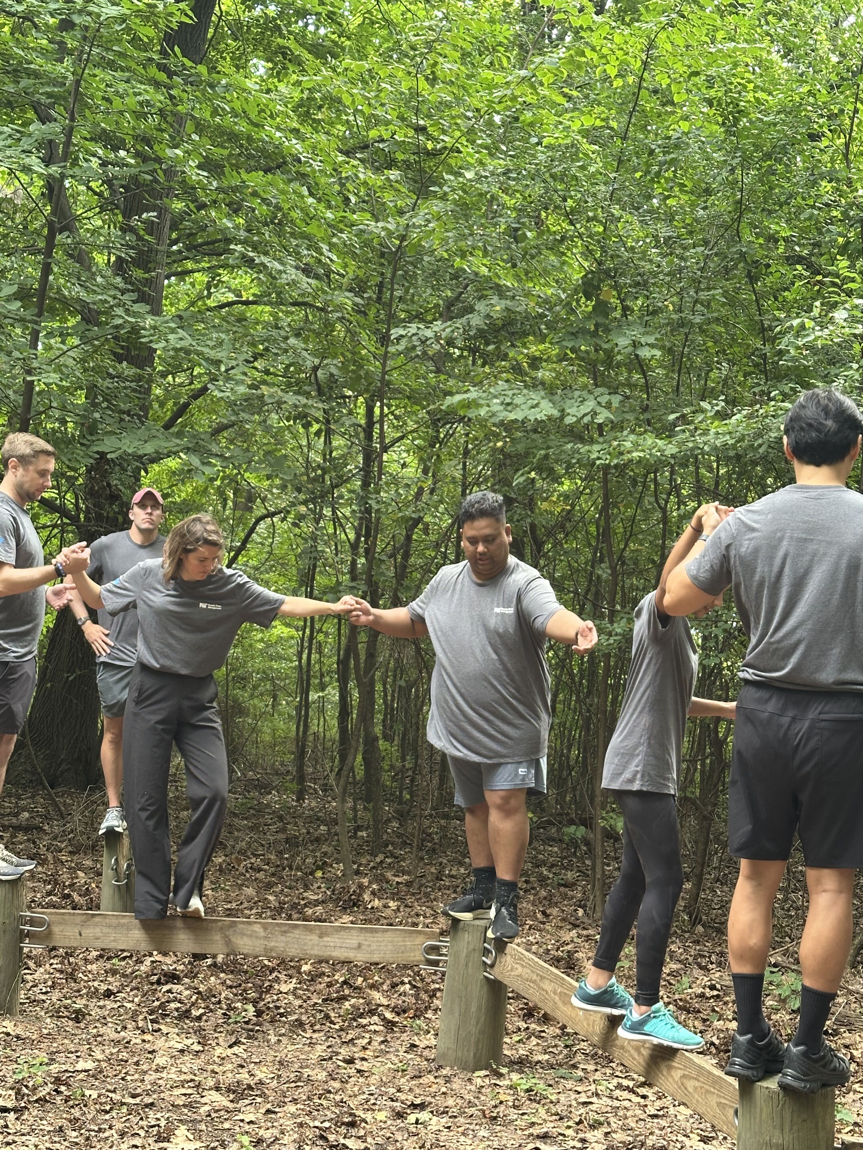A group of people are participating in an outdoor team-building activity in a wooded area. They are carefully walking across a series of wooden beams elevated by posts, holding onto each other for support and balance. The participants are dressed in matching gray t-shirts and shorts, suggesting a coordinated event or company outing. The activity appears to be a challenge that requires cooperation and trust among the team members as they navigate the obstacle course.